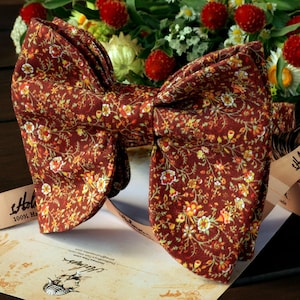 Peut inclure: Un nœud papillon bordeaux orné d'un motif floral jaune, orange et blanc. Le nœud papillon est centré sur un papier de style vintage avec un arrangement floral en arrière-plan. Un ruban avec du texte est également visible.