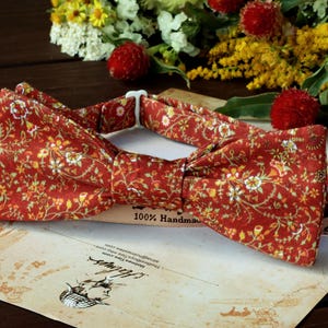 May include: A red bow tie with a floral pattern in shades of yellow and white. The bow tie is handmade and has a label that says "100% Handmade".