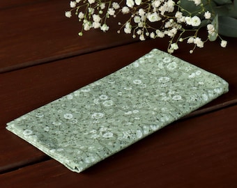 Può includere: Un fazzoletto di stoffa verde con un motivo floreale bianco. Il fazzoletto è piegato e appoggiato su una superficie di legno.