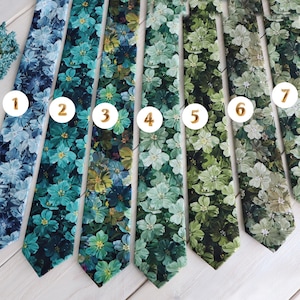 Op de afbeelding: Zeven stropdassen met bloemenpatroon in blauw- en groentinten worden getoond. Elke stropdas heeft een ander bloemmotief, met variaties in kleur en patroon. De stropdassen zijn naast elkaar gerangschikt en tonen de verscheidenheid aan ontwerpen.