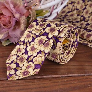 May include: A purple tie with a white floral pattern. The tie is rolled up and has a gold trim.