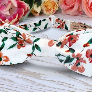 May include: White bow tie with a floral pattern featuring pink flowers and green leaves. The bow tie is handmade and has a label that says "100% handmade".