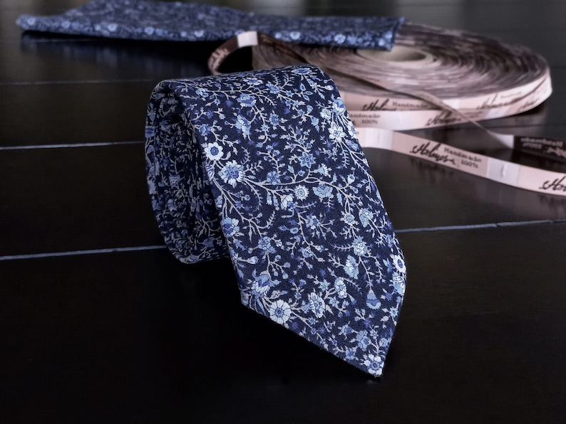 May include: A blue and white floral patterned tie with a rolled up portion of the tie visible. The tie is on a black surface with a brown and white label that says "Handmade 100%" and "Hobday's".