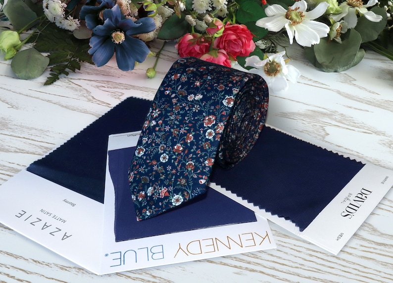 May include: A navy blue floral patterned tie with a white background. The tie is rolled up and displayed on a white wooden surface with two fabric swatches labeled "Kennedy Blue" and "AZZALE Matte Satin".