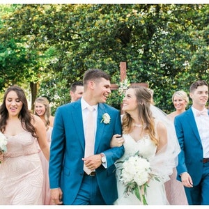 May include: A wedding party, including a bride and groom, walks outdoors. The groom wears a blue suit with a pink tie, and the bride wears a white dress and veil. Bridesmaids wear pink dresses, and groomsmen wear blue suits with pink bow ties.
