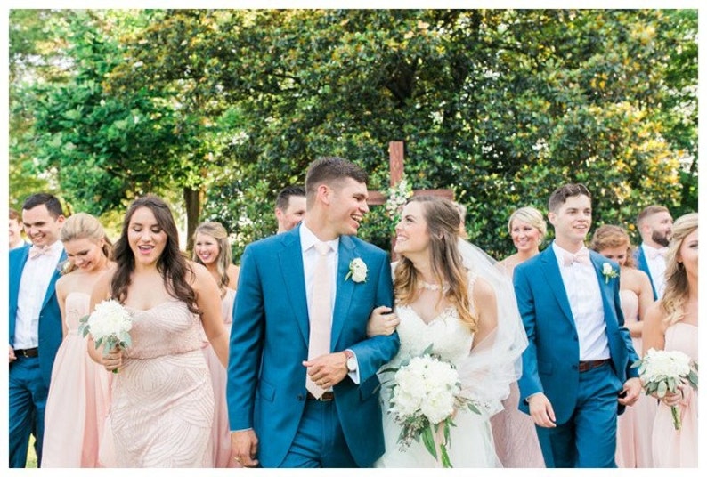 May include: A wedding party, including a bride and groom, walks outdoors. The groom wears a blue suit with a pink tie, and the bride wears a white dress and veil. Bridesmaids wear pink dresses, and groomsmen wear blue suits with pink bow ties.