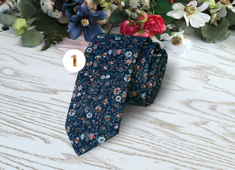 May include: A dark blue tie with a floral pattern featuring small white, pink, and orange flowers. The tie is rolled up and lying on a white wooden surface.