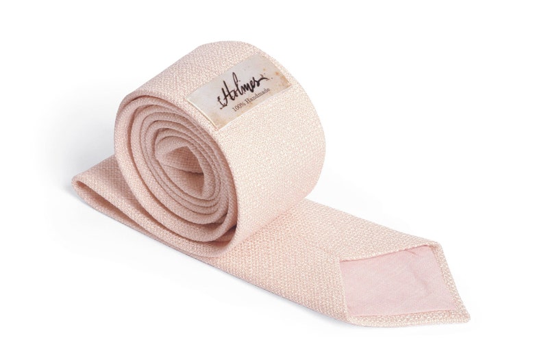 May include: A rolled-up, light pink necktie with a textured weave. The tie is unrolled at the end, revealing a lighter pink lining. A small label with the name "Halmer" and "100% Handmade" is attached to the rolled portion.