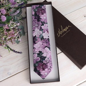 May include: A floral tie with a purple and white flower pattern, presented in a brown box. The tie is displayed in a white box, with the brand name "Holmes Classic Style" on the box lid. A bouquet of purple flowers is in the background.