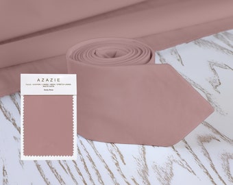 May include: A dusty rose colored tie made of matte satin fabric. The tie is rolled up and displayed on a white wooden surface. A fabric swatch is also shown with the text "A ZAZIE Tulle / Chiffon / Lining / Mesh / Stretch Lining Matte Satin Dusty Rose"