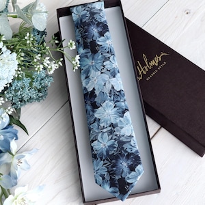 May include: A floral patterned tie in a presentation box. The tie features a blue and white floral design. The box is dark brown with the gold text "Holmes CLASSIC STYLE". A bouquet of blue and white flowers is in the background.