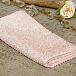 May include: A folded, light pink linen napkin with a textured weave. The napkin is placed on a rustic wooden surface, with a small white rose and pussy willow branches in the background. The napkin is folded neatly.
