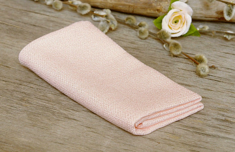 May include: A folded, light pink linen napkin with a textured weave. The napkin is placed on a rustic wooden surface, with a small white rose and pussy willow branches in the background. The napkin is folded neatly.