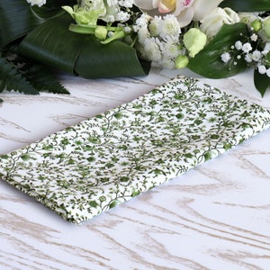 Op de afbeelding: Een witte stoffen servet met een groen bloemenpatroon. De servet is gevouwen en ligt op een wit houten oppervlak.