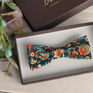Op de afbeelding: Een marineblauwe vlinderdas met een bloemenpatroon in oranje, teal en geel, gepresenteerd in een bruine doos met een witte binnenkant. De vlinderdas is omgeven door groen en bloemen.