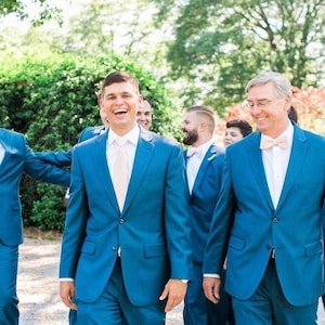 May include: A group of people in teal suits with white shirts and various colored ties and bow ties. The group is walking outside on a sunny day. The suits are a solid color and appear to be made of a smooth fabric.