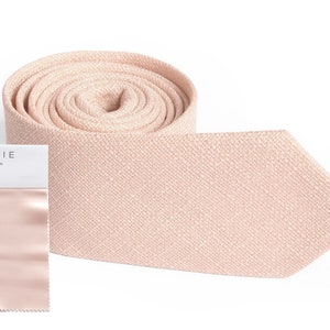 May include: A rolled-up, light pink matte satin tie with a matching swatch and packaging. The packaging reads "AZAZIE MATTE SATIN Pearl Pink". The tie is a solid color and has a textured appearance.