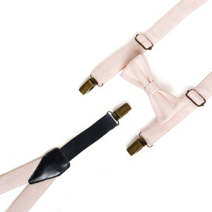 May include: A light pink bow tie and suspenders set with brass-colored metal hardware. The suspenders are light pink with black leather detailing and black clips. The bow tie is also light pink.