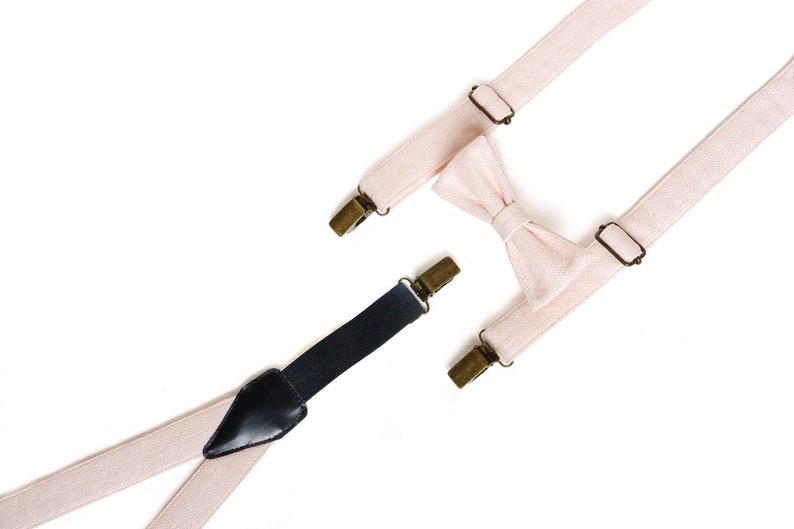 May include: A light pink bow tie and suspenders set with brass-colored metal hardware. The suspenders are light pink with black leather detailing and black clips. The bow tie is also light pink.