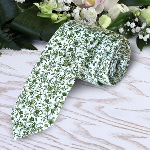Op de afbeelding: Een witte stropdas met een groen bloemenpatroon. De stropdas is opgerold en ligt op een wit houten oppervlak.