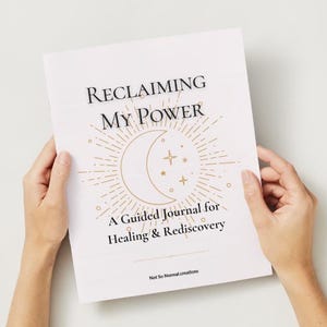 Op de afbeelding: Een wit tijdschrift getiteld "Reclaiming My Power" met gouden hemelse illustraties en tekst. Het tijdschrift wordt door twee handen tegen een witte achtergrond gehouden. Het tijdschrift is een geleid tijdschrift voor genezing en herontdekking.