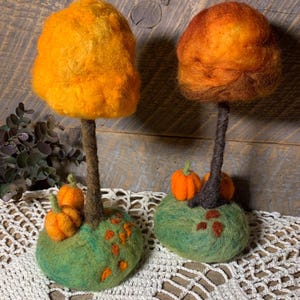 May include: Two needle-felted tree sculptures with vibrant orange and red wool foliage. Each tree has a dark brown trunk rooted in a rounded green base. The bases are decorated with small orange felt pumpkins and scattered brown leaf accents.