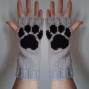 Peut inclure: Une paire de mitaines grises tricotées, chacune ornée d'une grande empreinte de patte noire. Les gants sont faits d'un fil doux et texturé, conçus pour garder les mains au chaud tout en laissant les doigts libres.