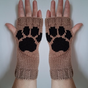 Könnte beinhalten: Beige gestrickte Fingerhandschuhe mit einem schwarzen Pfotenabdruck-Design auf jeder Handfläche. Die Handschuhe sind aus einem strukturierten Garn gefertigt und bedecken die Hand bis zu den Knöcheln. Ein warmes Accessoire.