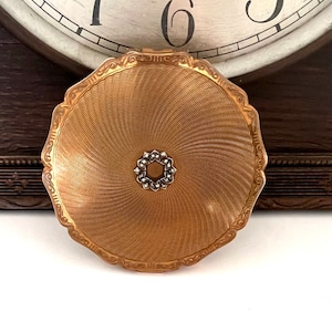 Può includere: Un compatto dorato con un design a spirale e un decoro centrale di strass.