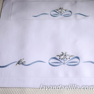 May include: Two white linen table runners with blue embroidered ribbon and floral designs. The bottom runner has a larger bow and the text "lavandaelilla.com" is embroidered on the bottom right corner.