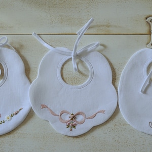 May include: Three white baby bibs with embroidered designs. The first bib has a pink butterfly and flowers, the second has a pink bow, and the third has two cats with hearts.