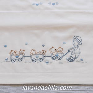 Puede incluir: Ropa de cama color crema con detalles bordados en azul. Un niño tira de un tren de vagones llenos de ositos de peluche. Corazones azules y detalles florales salpican la tela. El sitio web lavandaelilla.com es visible.