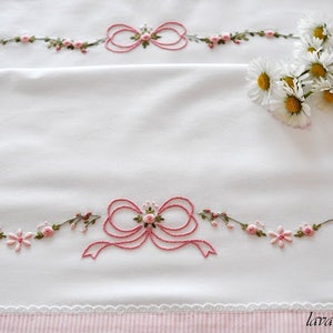 Può includere: Biancheria da letto bianca con fiocchi ricamati rosa e dettagli floreali. La biancheria da letto ha un bordo a righe rosa e bianco.