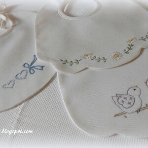 May include: Two white baby bibs with embroidered designs. One bib has a row of blue hearts and bows. The other bib has a row of white flowers and two birds on a branch.