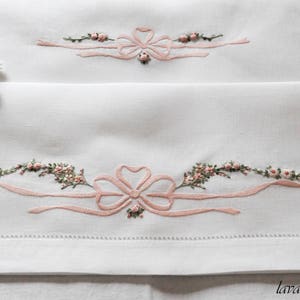 Puede incluir: Dos fundas de almohada blancas con delicados diseños florales bordados en rosa y lazos. El bordado presenta pequeñas flores rosas y hojas, creando un aspecto delicado y femenino.