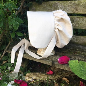 May include: A white linen bonnet with a gathered ruffle around the crown and a long, tied ribbon. The bonnet is sitting on a wooden bench with rose petals scattered around it.