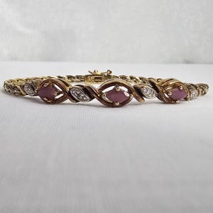 May include: A gold-toned bracelet featuring alternating oval-shaped ruby gemstones and diamond accents. The bracelet has a woven chain design and a secure clasp. The gemstones are a deep red color, and the diamonds sparkle.