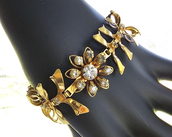 Puede incluir: Pulsera dorada con diseños florales y lazos. La pulsera tiene una flor central con un gran brillante transparente rodeado de brillantes más pequeños. Los lazos y las flores están conectados por eslabones dorados. La pulsera se muestra sobre una superficie negra.