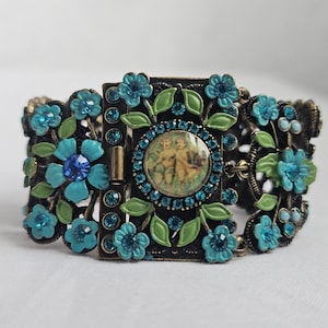 Puede incluir: Una pulsera ornamentada de estilo antiguo con acabado bronce. Presenta flores azul turquesa, hojas verdes y cristales azules brillantes. El centro tiene un camafeo circular con querubines.