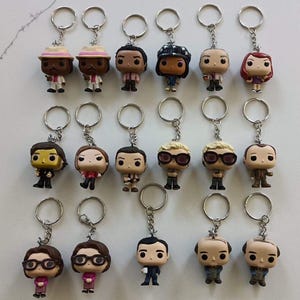 May include: A collection of Funko Pop-style keychains featuring various character designs. Each keychain has a silver metal ring and chain. The figures have detailed features, including clothing and hairstyles. The keychains are arranged on a white surface.