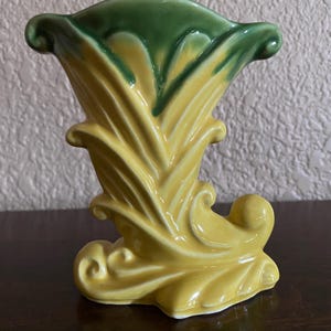 Könnte beinhalten: Eine Keramikvase mit einer glänzend gelben Basis, skulpturalen blattartigen Mustern und einem dunkelgrün glasierten Rand. Das Gefäß hat eine ausgestellte Öffnung und eine geschwungene, stiefelartige Silhouette mit detaillierten Verzierungen.