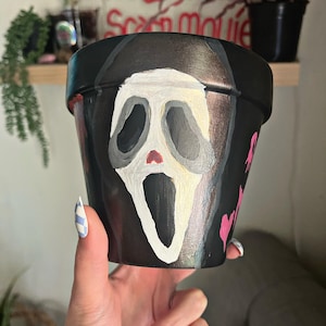 May include: Hand-painted black flower pot featuring the Ghostface mask from the Scream movie franchise. The white mask has a red mouth and is set against a black background. The pot is held by a hand with blue and white checkered nails.