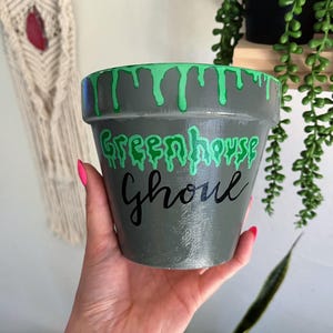 May include: Gray painted ceramic plant pot with green dripping paint around the rim. The words "Greenhouse Ghoul" are written in green and black. A decorative item for plants.