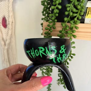 May include: Black ceramic planter with the words "THORNS & THR" in neon green. The planter sits on a black metal stand. A hand with pink nail polish holds the stand. A shelf with a black pot and green plants is in the background.