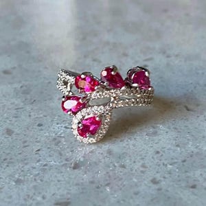 May include: A silver ring featuring a cluster of vibrant pink gemstones and small clear stones. The ring has a flowing, organic design, with the pink stones arranged in a decorative pattern.