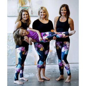 May include: Three women are wearing black tank tops and colorful leggings with a watercolor-like print. One woman is holding a young girl in the air, who is also wearing a purple tank top and colorful leggings.
