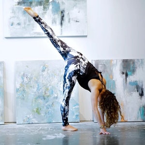 May include: A woman in black and white patterned leggings performs a yoga pose in front of abstract paintings. Her leg is extended high in the air, and she is bending forward at the waist.