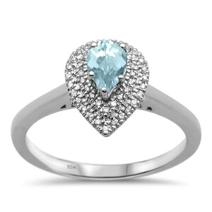 Könnte beinhalten: Ein silberner Ring mit einem tropfenförmigen, hellblauen Edelstein, umgeben von kleinen, klaren Steinen. Der Ring hat ein zartes Band und ein Halo-Design. Dies ist ein Schmuckstück.