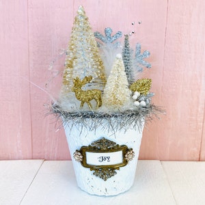 May include: White ceramic planter with a silver glitter rim, filled with white and gold glitter Christmas trees, a gold glitter reindeer, and a gold sign that reads "Joy".