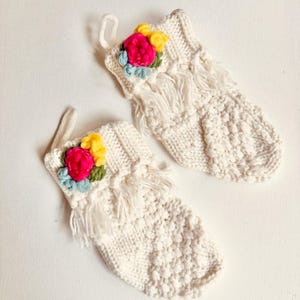 Puede incluir: Dos calcetines de Navidad de punto color crema con adornos florales coloridos. Cada calcetín presenta un patrón de punto texturizado, detalles de flecos y un lazo para colgar. Las flores son rosas, amarillas y azules.